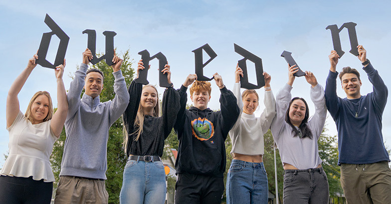 Seven smiling young people hold up letters spelling DUNEDIN.