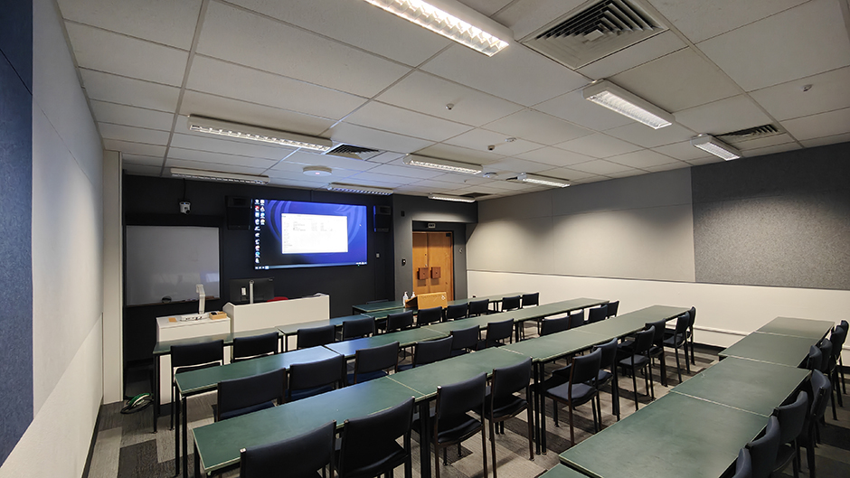 View of Burns 4 seminar room lefthand from back, with tables and chairs in rows facing a projector screen