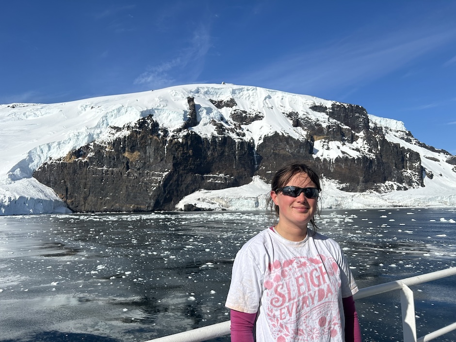 A young woman in sunglasses and t-shirt stands on the deck of a ship; behind her is water dotted with ice, a rocky cliff swathed with snow and ice, and bright blue sky.