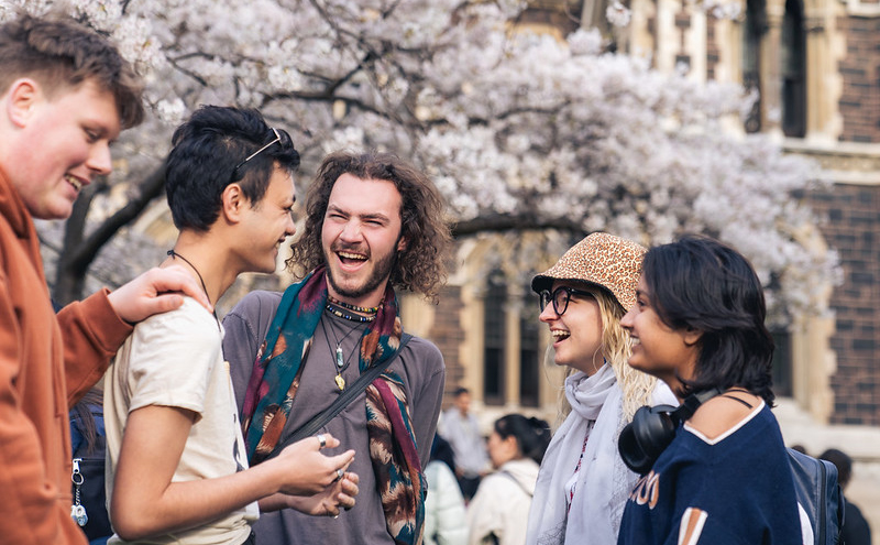 A group of five young people talking and laughing, with a historic building and blossom-covered tree in the background.