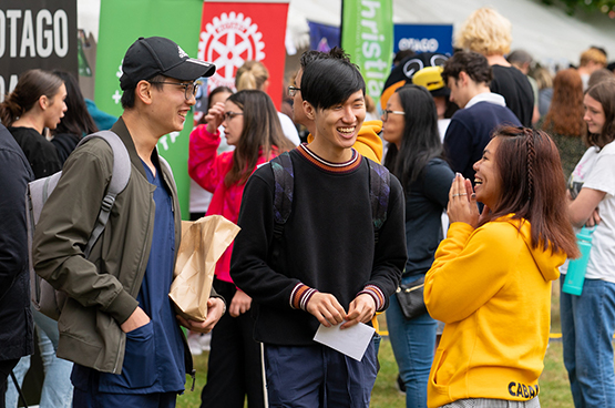 Three young people stand talking and laughing in a grassy outdoor area, surrounded by a crowd of young people and banners on display. 