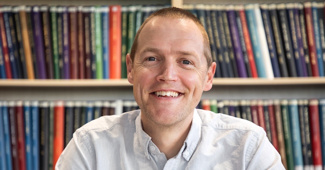 Prof Peter Mace smiles at the camera with a bookshelf behind him.