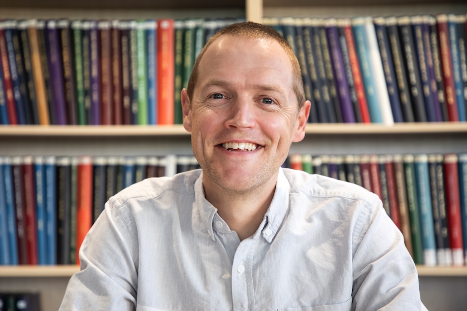 Prof Peter Mace smiles at the camera with a bookshelf behind him.