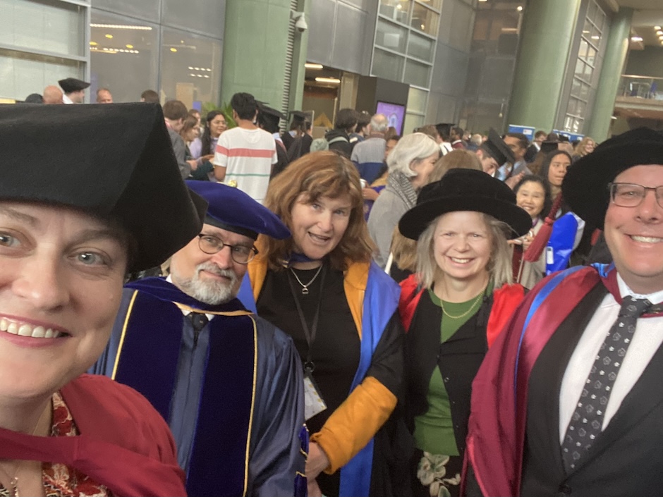 A group of people dressed in academic robes grinning at the camera in front of a crowd of people.