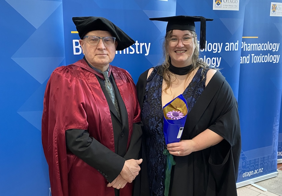 Two people dressed in academic robes pose for a photo in front of blue banners.