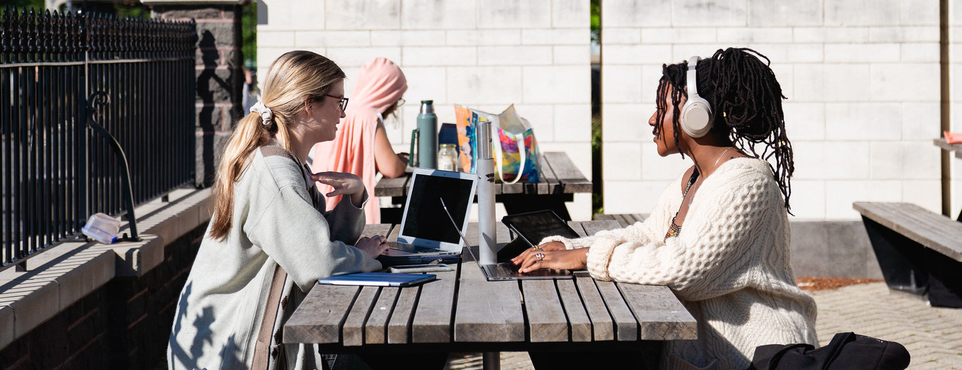 Two students with laptops out at an outdoor table.