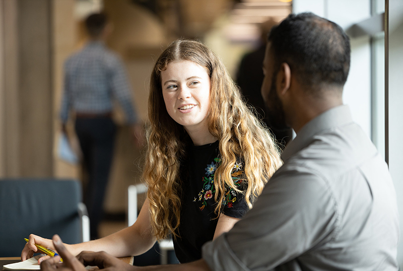 A young man and woman sit side by side at a table talking and working together