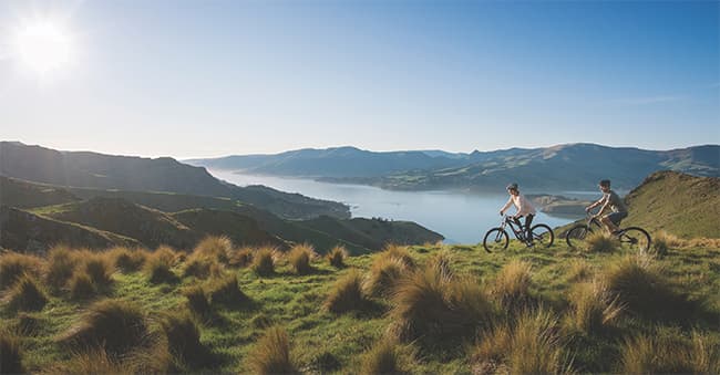 Two people ride bicycles through a grassy high-country area, with a harbour and hills in the background.