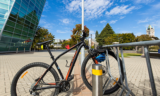 A bicycle parked in a bike rack in a paved area, with the University of Otago Clocktower visible in the background