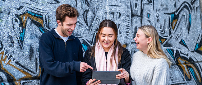 Three students standing in front of a mural, looking at a tablet and laughing together