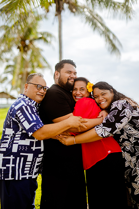 An aiga of four Samoan adults in a group hug, with palm trees visible in the background