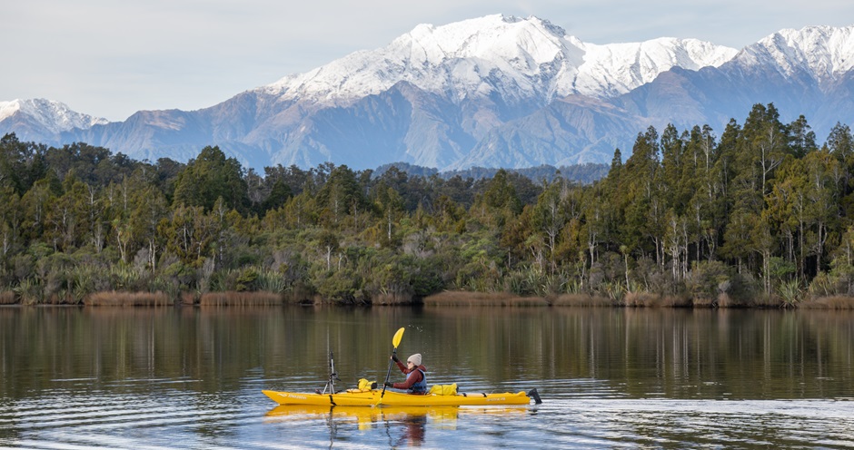 A person kayaking on Ōkārito Lagoon, a still expanse of water surrounded by forest, with mountains visible in the distance.