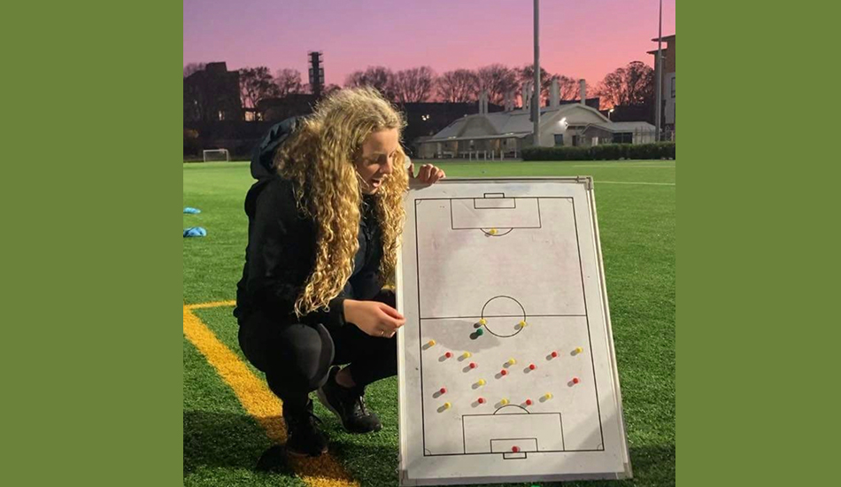 Woman with curly blonde hair on a football pitch at night with a whiteboard planning game strategies.