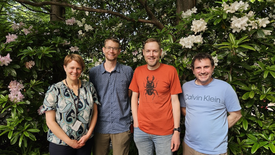 Four people stand in front of flowering rhododendron bushes.