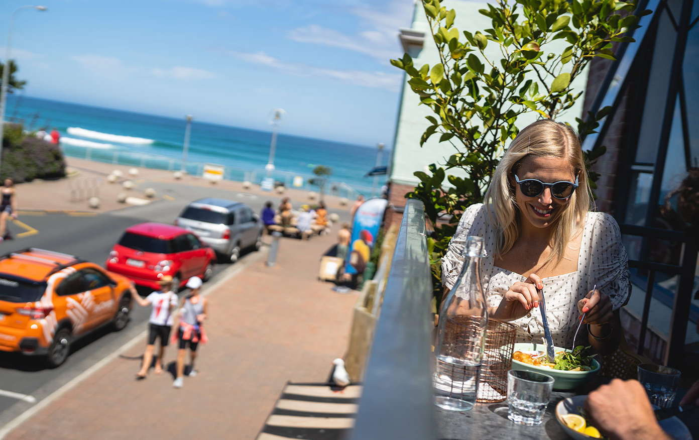 People eating on a balcony in the sun, with St Clair Esplanade and the ocean in the background.