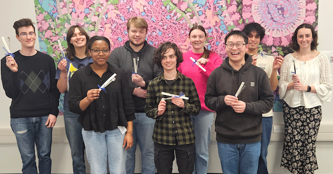 A group of standing, smiling students hold up little metal spatulas.